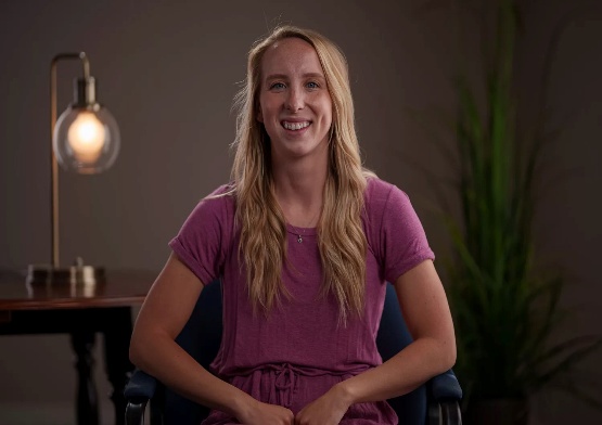 A photo of Kendra Brown, a young white woman with long blonde hair almost to her waist. She is smiling at the camera in a purple short-sleeve top with a lamp in the background.