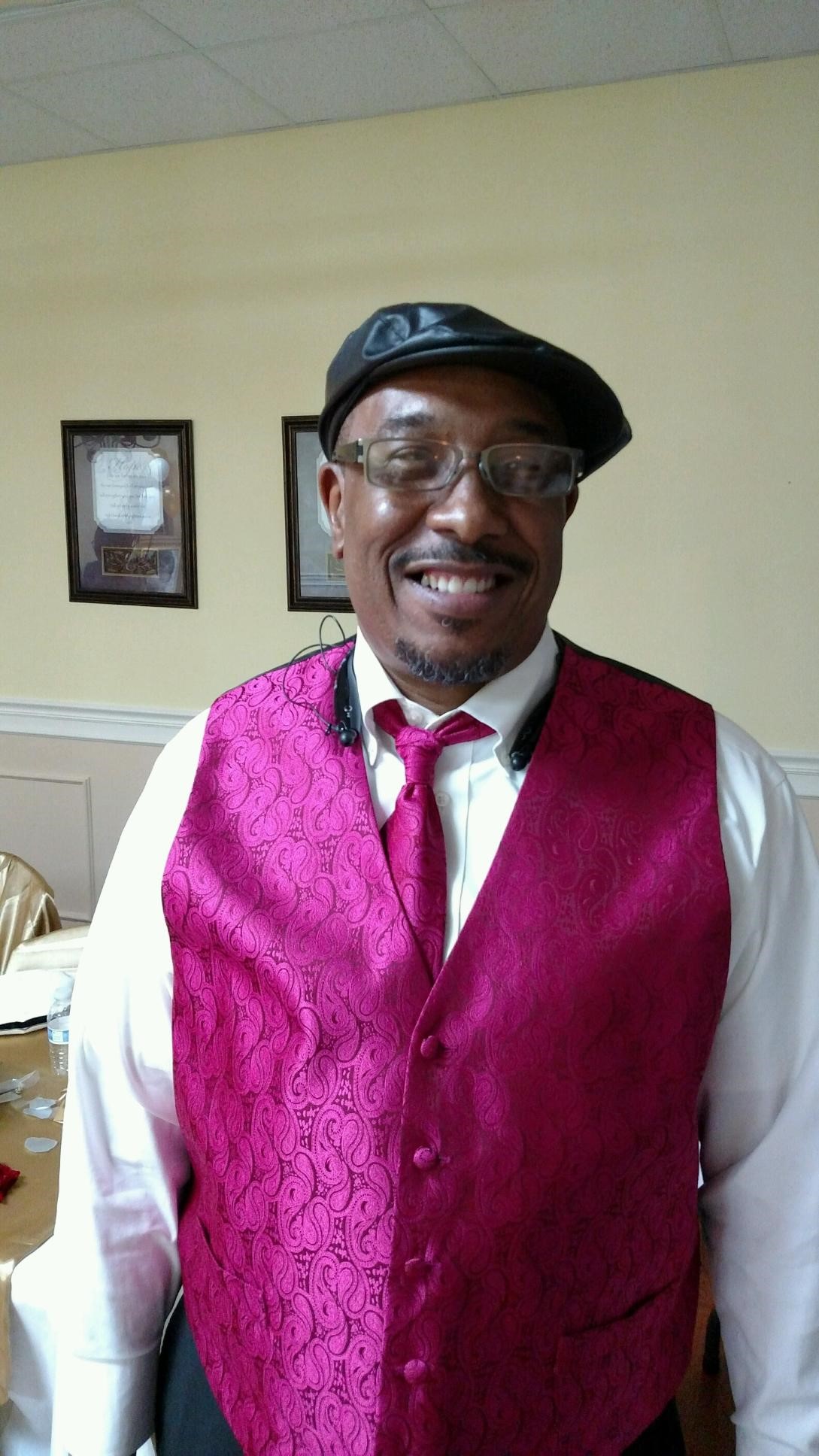 Black Man smiling big with a magenta vest, long sleeved white shirt and a hat