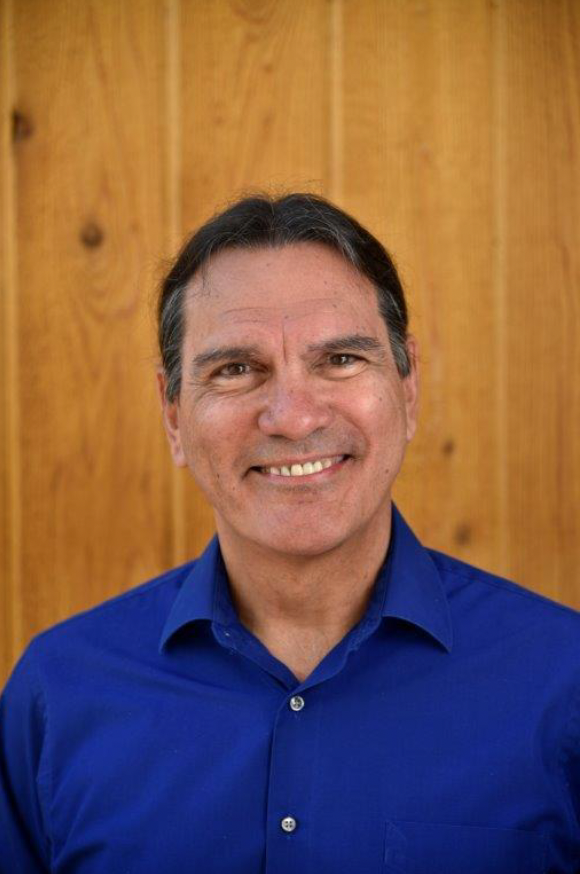 Photo of Andrew Johnson, a Cherokee man with short dark hair. He is smiling at the camera with a bright blue button up shirt on standing in front of a wood panel wall.