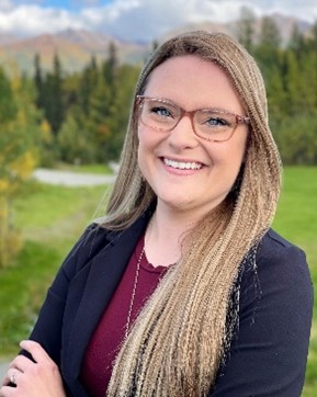 Photo of a young white woman with long blonde hair past her shoulders and glasses. She is posing for a photo outside.