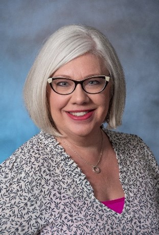 Photo of a white woman with chin length gray hair, bright lipstick, and dark rimmed glasses. She is posing for a photo in front of a blue background.