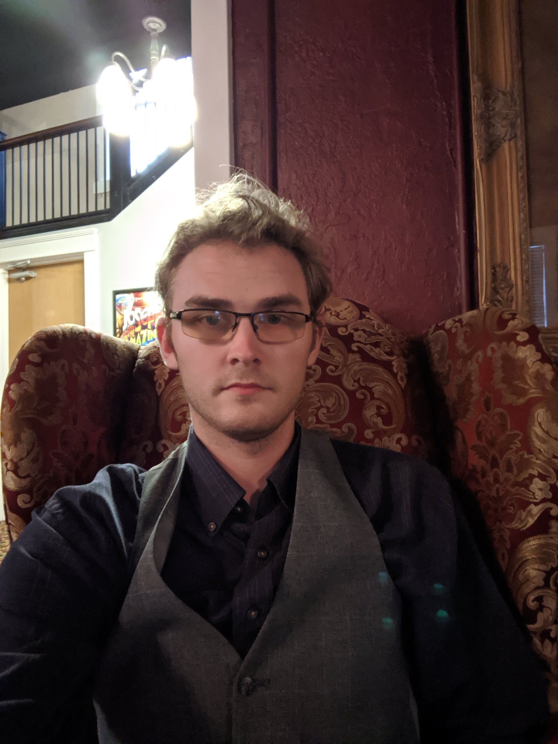 Photo of Noah Russell, a young white man with glasses and dark hair. He is sitting in a chair in formal attire.