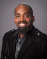 A Black male with a beard and mustache smiling at the camera. He is wearing a dark colored suit with a tie.