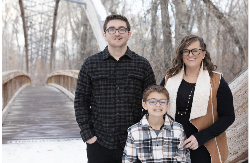 Photo of Brittany Shipley, a young white woman with long, wavy brown hair and dark rimmed glasses standing beside her two sons. One son is taller than her and the other comes up to her shoulders. Both also wear glasses. All are standing outside in the snow, posing for a family photo.