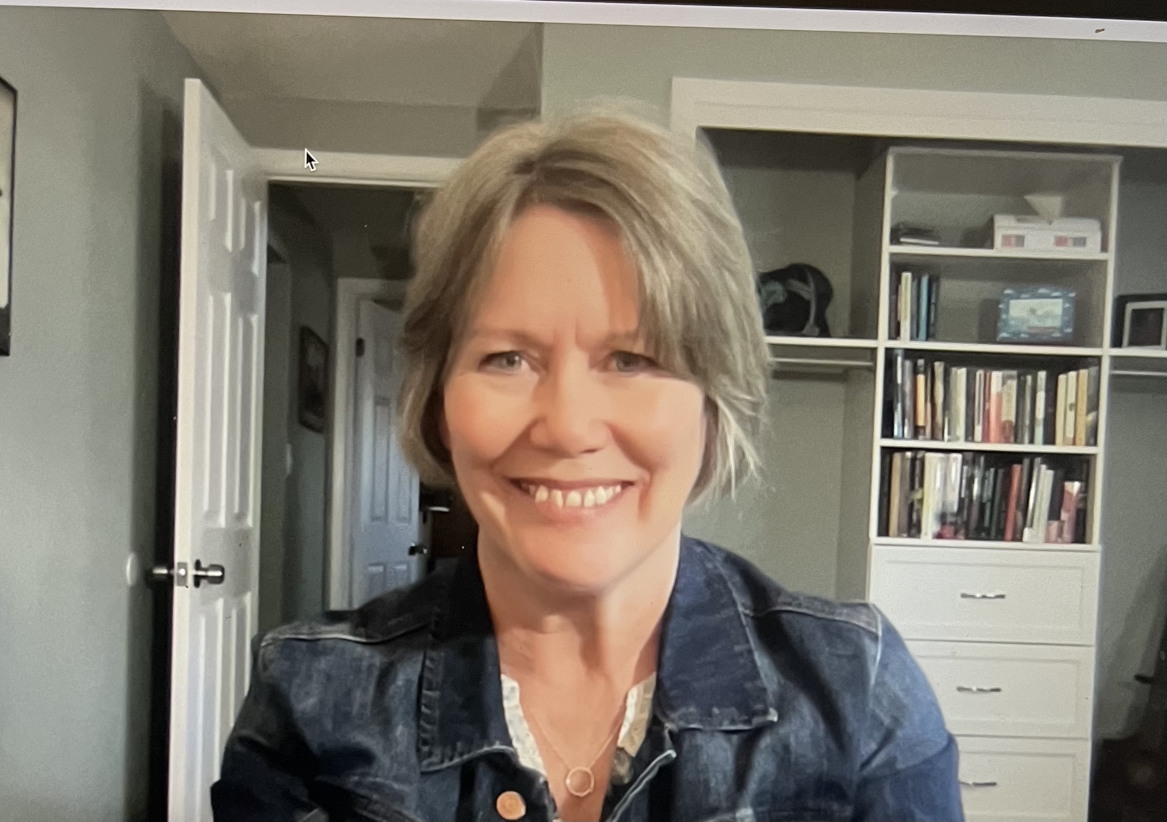 A photo of Rebecca Roberts, a white woman with ash blonde hair smiling for a photo. A bookcase is behind her and she is wearing a jean jacket.