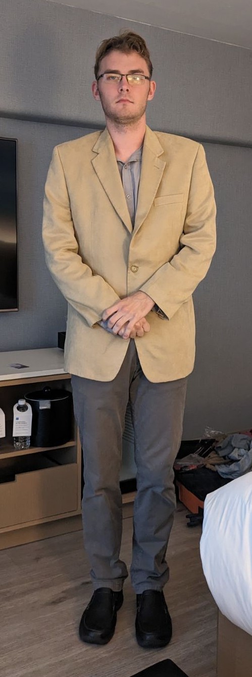 Full body photo of Noah Russell. Noah is a young white man with shorter brown hair and glasses. He is wearing a tan color suit.